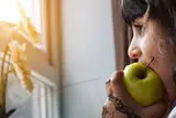 Kind beißt in einen Apfel und schaut aus dem Fenster